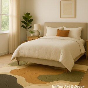 May include: A bedroom scene featuring a bed with cream-colored bedding and an orange accent pillow. A patterned rug with abstract shapes in shades of green, brown, and beige is on the floor. The text "Indian Art & Decor" is visible on the rug.