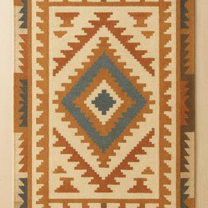 May include: Rectangular beige rug with a geometric pattern in shades of brown and blue. The rug features a central diamond design and a border with repeating shapes. The text "Indian Art & Decor" is visible at the bottom.
