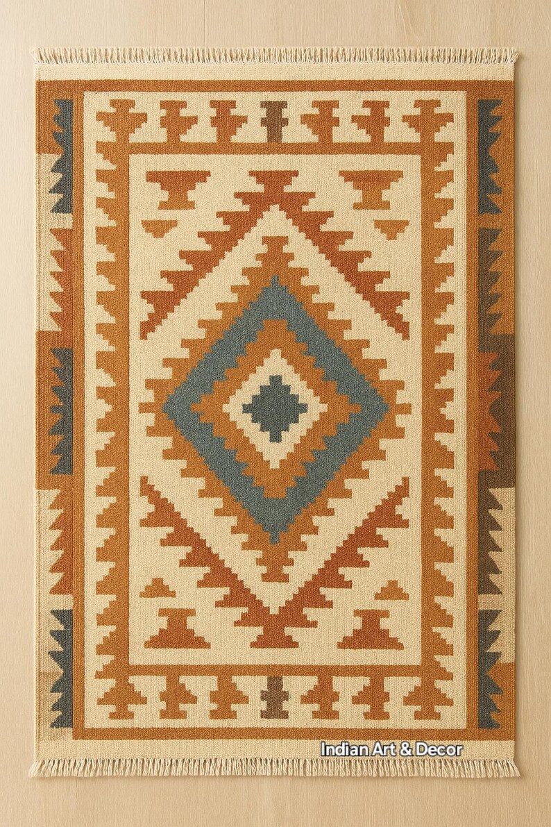 May include: Rectangular beige rug with a geometric pattern in shades of brown and blue. The rug features a central diamond design and a border with repeating shapes. The text "Indian Art & Decor" is visible at the bottom.