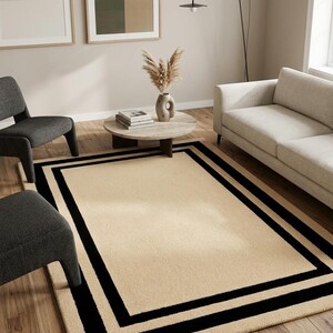 May include: A rectangular area rug featuring a solid beige center with a double black border frame. The rug has a textured surface and a small corner label that reads Hand Tufted in India. The design is flat and covers a wooden floor area.