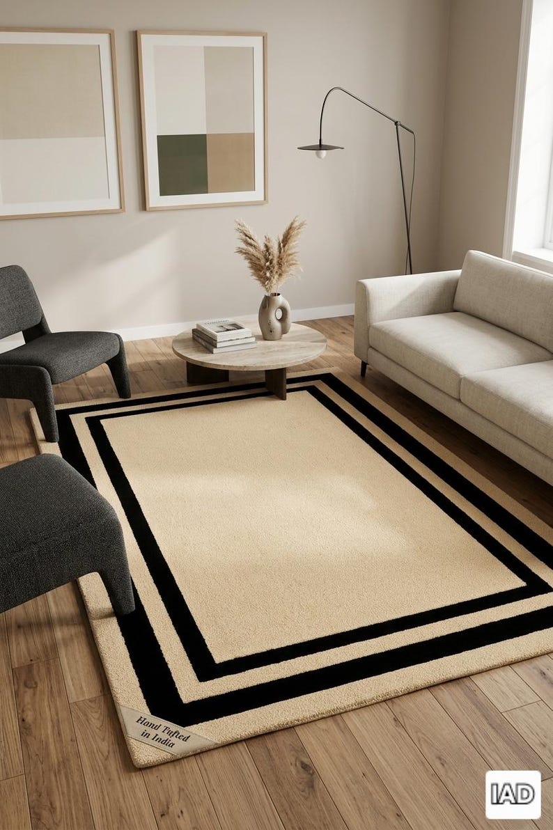 May include: A rectangular area rug featuring a solid beige center with a double black border frame. The rug has a textured surface and a small corner label that reads Hand Tufted in India. The design is flat and covers a wooden floor area.