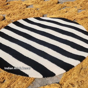 Op de afbeelding: Een ronde vloerkleed met opvallende zwart-witte strepen. Het ontwerp van het vloerkleed is opvallend en geschikt voor moderne interieurs. Bundels goudkleurig garen omringen het vloerkleed.