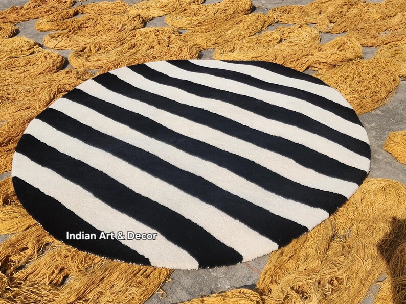 Op de afbeelding: Een ronde vloerkleed met opvallende zwart-witte strepen. Het ontwerp van het vloerkleed is opvallend en geschikt voor moderne interieurs. Bundels goudkleurig garen omringen het vloerkleed.