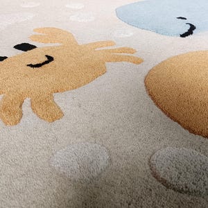 May include: A beige area rug featuring a tufted design of a tan crab with black eyes and a smile, a light blue whale, and a large tan circle. The rug surface includes scattered white circular shapes and a textured pile throughout the fabric.