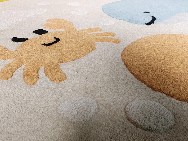 May include: A beige area rug featuring a tufted design of a tan crab with black eyes and a smile, a light blue whale, and a large tan circle. The rug surface includes scattered white circular shapes and a textured pile throughout the fabric.