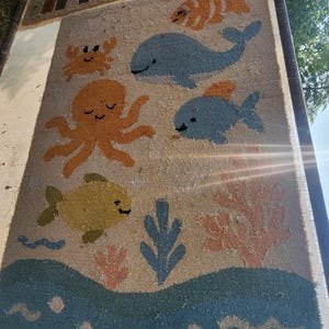 May include: A rectangular coir doormat featuring a tan background with a colorful marine life pattern. The design includes a blue whale, an orange octopus, a crab, and various fish in shades of blue, yellow, and orange, alongside coral and wave motifs.