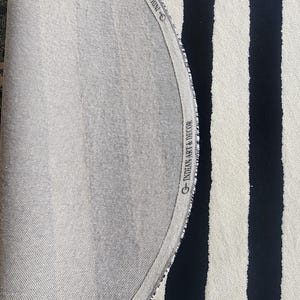 Op de afbeelding: Een gedeelte van een gestreept tapijt met afwisselend zwarte en cr&egrave;mekleurige verticale strepen. De rand van het tapijt toont een lichtgrijze achterkant met de tekst "INDIAN ART & DECOR" langs de rand.