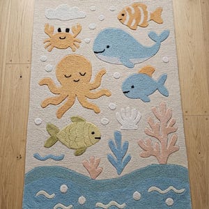 May include: A rectangular area rug with a cream background featuring a tufted ocean theme. The design includes a blue whale, a yellow octopus, a crab, various fish, coral, and a shell. The bottom section depicts blue waves with white bubbles.