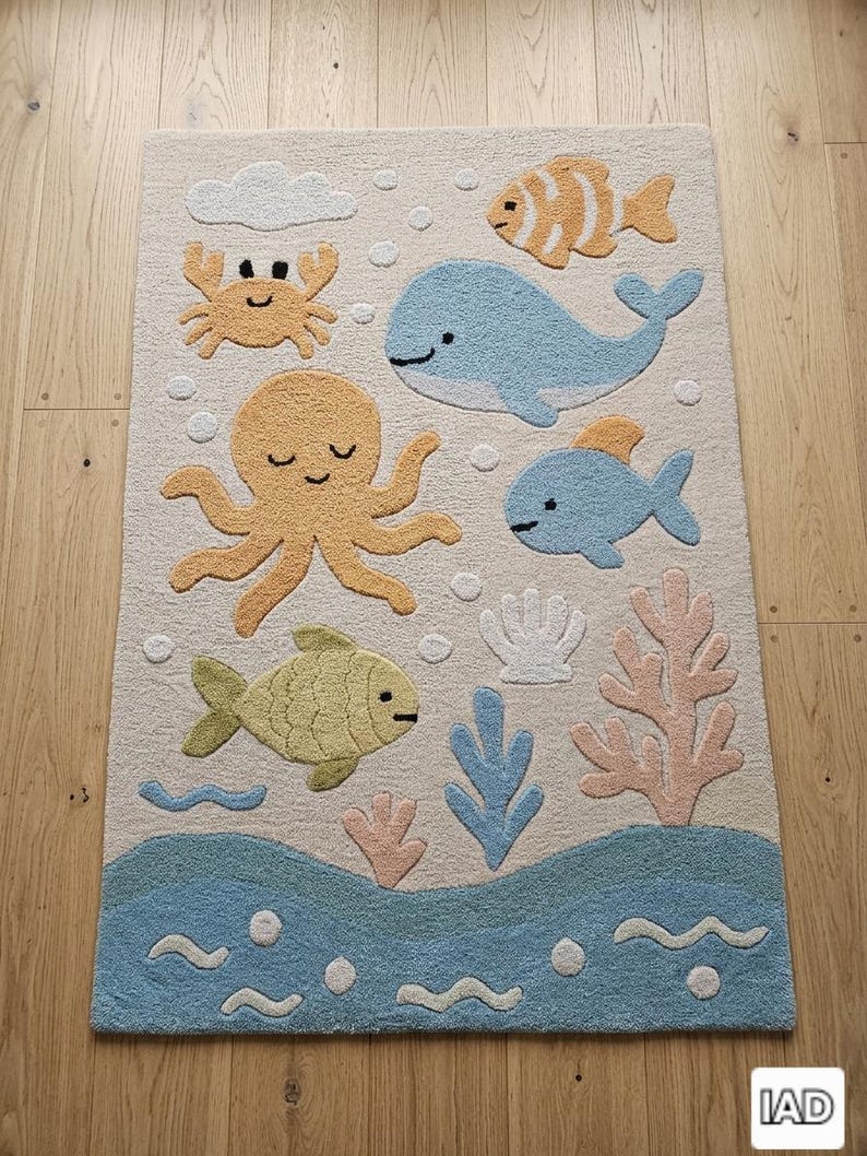 May include: A rectangular area rug with a cream background featuring a tufted ocean theme. The design includes a blue whale, a yellow octopus, a crab, various fish, coral, and a shell. The bottom section depicts blue waves with white bubbles.