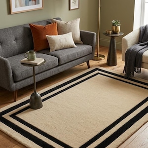 May include: A rectangular area rug featuring a solid beige center bordered by two distinct black rectangular frames. The rug has a textured pile surface and is positioned on a wooden floor. The letters IAD are visible in the bottom right corner.