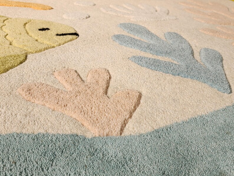 May include: A tufted area rug featuring a cream-colored background with raised, textured motifs of a yellow fish, light blue coral, and peach-colored coral. The design includes soft, muted tones and a plush, high-pile surface texture throughout.