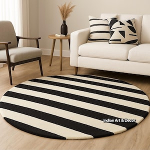 Op de afbeelding: Een ronde vloerkleed met brede horizontale strepen in zwart en cr&egrave;me. Het kleed ligt in een woonkamer, voor een witte bank, sierkussens en een houten bijzettafel. Het ontwerp van het kleed geeft de ruimte een moderne uitstraling.