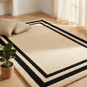 May include: A rectangular area rug featuring a solid beige center with a double black border frame. The rug has a textured, plush pile surface and is positioned on a light-colored wooden floor. The text IAD is visible in the bottom right corner.