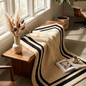 May include: A beige textured throw blanket featuring two bold black stripes along its border, draped over a wooden bench. The blanket has a soft, plush appearance and is positioned near a ceramic vase with dried grasses and an open book.