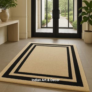May include: A rectangular area rug featuring a solid beige center framed by a thick black border. The rug has a textured surface and includes the white text Indian Art & Decor printed on the lower section of the black border area.