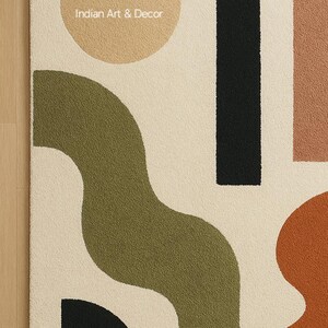 K&ouml;nnte beinhalten: Cremefarbener Teppich mit einem abstrakten Design mit einem beigen Kreis, einem schwarzen Rechteck und olivgr&uuml;nen und rostfarbenen organischen Formen. Der Text "Indian Art & Decor" ist auf dem Teppich zu sehen.