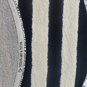 Op de afbeelding: Een ronde vloerkleed met afwisselende zwarte en cr&egrave;mekleurige verticale strepen. De rand van het vloerkleed heeft een grijze stoffen rand met de tekst "INDIAN ART & DECOR". Het vloerkleed heeft een getextureerd oppervlak.