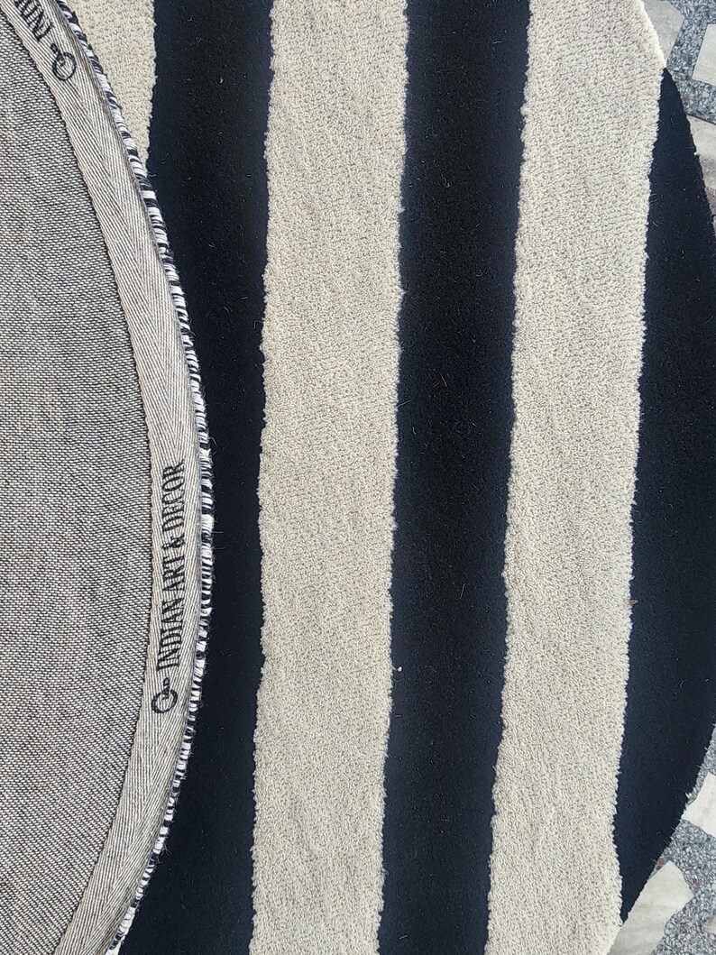 Op de afbeelding: Een ronde vloerkleed met afwisselende zwarte en cr&egrave;mekleurige verticale strepen. De rand van het vloerkleed heeft een grijze stoffen rand met de tekst "INDIAN ART & DECOR". Het vloerkleed heeft een getextureerd oppervlak.