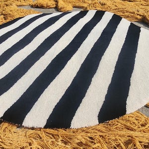Op de afbeelding: Een ronde vloerkleed met afwisselende zwarte en witte strepen. Het kleed heeft een zacht, getextureerd uiterlijk. Gele garenstrengen omringen het kleed, wat wijst op een handgemaakt of ambachtelijk product.