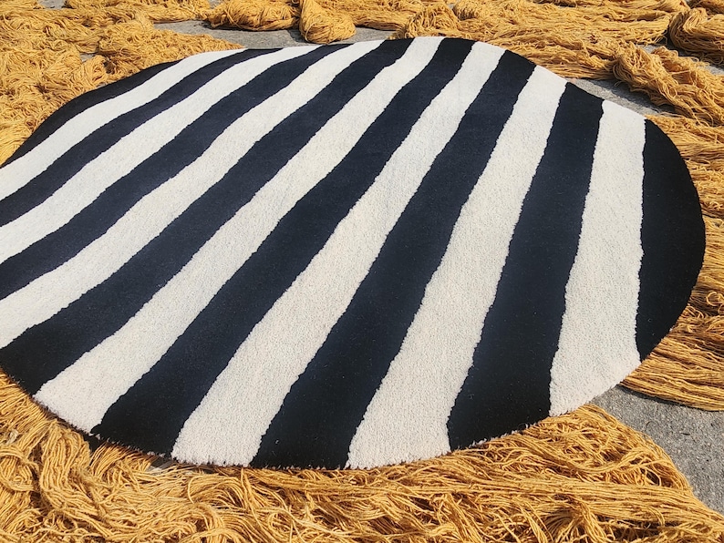 Op de afbeelding: Een ronde vloerkleed met afwisselende zwarte en witte strepen. Het kleed heeft een zacht, getextureerd uiterlijk. Gele garenstrengen omringen het kleed, wat wijst op een handgemaakt of ambachtelijk product.
