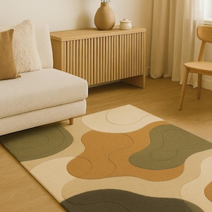 May include: A modern area rug with an abstract design in shades of beige, brown, and olive green. The rug features a wavy, organic pattern and is placed in a living room setting with a sofa, wooden cabinet, and chair. The text "Indian Art & Decor" is visible.