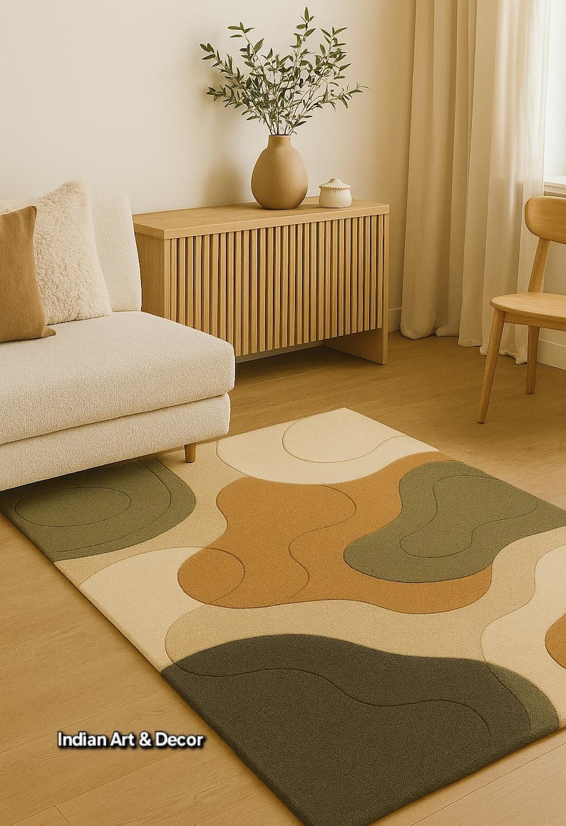May include: A modern area rug with an abstract design in shades of beige, brown, and olive green. The rug features a wavy, organic pattern and is placed in a living room setting with a sofa, wooden cabinet, and chair. The text "Indian Art & Decor" is visible.