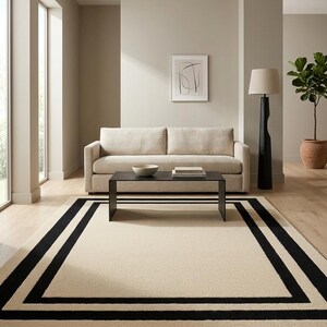 May include: A rectangular area rug featuring a large cream-colored center field framed by two distinct, thick black border lines. The rug is positioned on a light-toned wooden floor in a room with a beige sofa, a black coffee table, and a floor lamp.