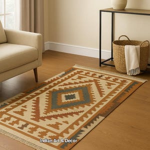 May include: Rectangular area rug with a geometric pattern in beige, brown, and teal. The rug has a fringed edge and is placed on a hardwood floor. The text "Indian Art & Decor" is visible.