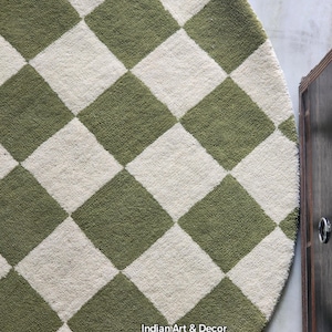 May include: A round area rug featuring a repeating checkered pattern in olive green and off-white. The rug has a textured, tufted surface and includes the text Indian Art & Decor printed in small white letters near the bottom edge of the rug.