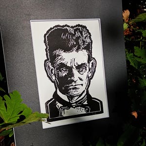 May include: A black and white linocut print of a man's portrait. The man has short, dark hair, a stern expression, and is wearing a suit with a bow tie. The print is mounted on a white mat and a black background.