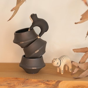 May include: A stack of three black ceramic bowls with a black cat figurine perched on top. A white and tan tiger figurine stands nearby. The objects are on a wooden surface with dried leaves in the background.