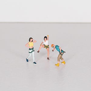 Puede incluir: Figuras en miniatura de tres personas con equipo de escalada. Una figura lleva una camiseta amarilla y pantalones blancos, otra una camiseta blanca y pantalones cortos, y la tercera una camisa azul y pantalones caqui. Todos posan como si estuvieran escalando.
