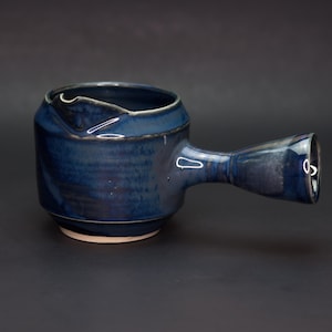 May include: A handmade, dark blue ceramic creamer with a handle. The creamer has a rounded body and a spout for pouring. The handle is a cylindrical shape. The creamer is approximately 6 inches long.