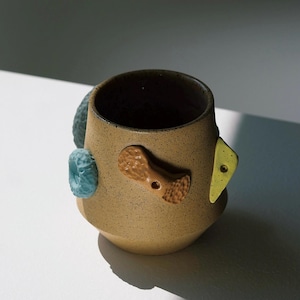 Könnte beinhalten: Eine braune Keramiktasse mit strukturierter Oberfläche und bunten Akzenten. Die Tasse hat ein gelbes, blaues und orangefarbenes Design.