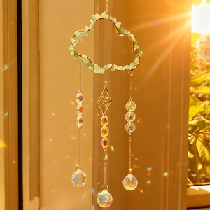 May include: A decorative suncatcher with a cloud-shaped frame adorned with green stones. Gold-coloured metal accents and hanging crystal beads create a prism effect. Designed to hang in a window.