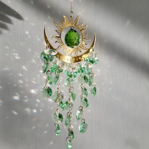 May include: A decorative suncatcher with a gold-coloured sun and crescent moon design. It features a large green crystal at the sun's centre, with hanging green crystal droplets. The suncatcher is suspended by a delicate chain.