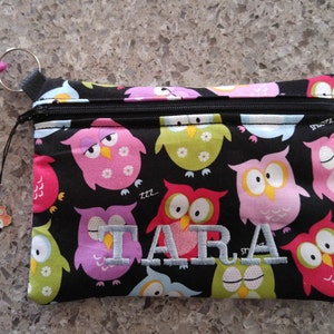 May include: A black zippered pouch with a colourful owl print. The pouch has a pink clip and a silver owl charm. The name "TARA" is embroidered on the front of the pouch.