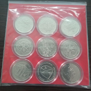 May include: Nine silver coins in clear plastic cases, arranged in a 3x3 grid. Each coin features a detailed design, with various symbols and text. The coins are displayed on a red background, inside a clear plastic bag.