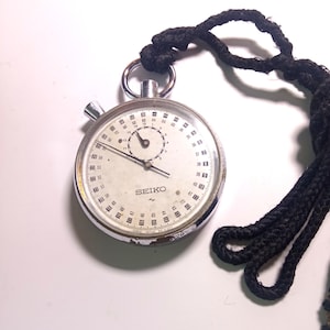 May include: A vintage Seiko stopwatch with a white face, black numerals, and a smaller inner dial. The stopwatch has a silver metal casing and a black cord attached for carrying. The watch shows signs of wear.