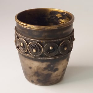 Puede incluir: Una taza pequeña de color plateado con una banda decorativa de círculos y puntos dorados alrededor del centro.