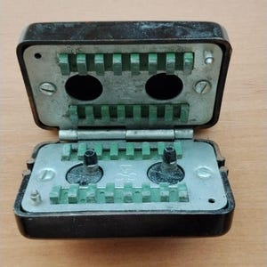 Puede incluir: Una caja de conexiones eléctrica rectangular de color marrón oscuro vintage con tapa abatible. El interior presenta dos placas metálicas con regletas de terminales de plástico verde y dos aberturas circulares. Dos pines negros visibles.