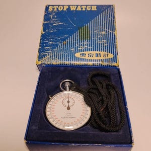 May include: A vintage stopwatch in a blue box. The watch face is white with black numerals and red markings. The box is labeled "STOP WATCH" and "TOKYO TOKEI". A black cord is attached to the stopwatch.