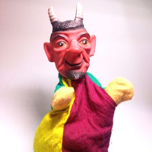 May include: A devil hand puppet with a red face, black beard, and horns. The puppet's clothing is a mix of yellow, green, and maroon fabric. The puppet has a smiling expression and is designed for hand operation.