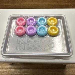 Pode incluir: Uma bandeja de plástico transparente contendo oito doces redondos e coloridos. Os doces são rosa, roxo, azul claro e amarelo. A bandeja tem a palavra "Kisses" impressa.
