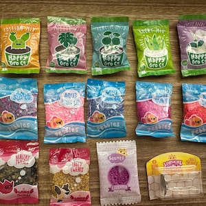 Puede incluir: Una colección de pequeñas bolsas de mezcla para macetas, guijarros y aromas para plantas. Las bolsas son de varios colores, incluyendo verde, rosa, azul y morado, con ilustraciones de plantas de dibujos animados y el texto "Happy Gro Co" y "Aqua Sun Co".
