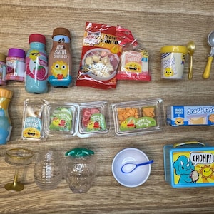 Pode incluir: Uma coleção de alimentos de brinquedo em miniatura, incluindo garrafas, recipientes e uma lancheira. Os itens estão dispostos em uma superfície de madeira. A paleta de cores inclui azul, amarelo, vermelho e verde.