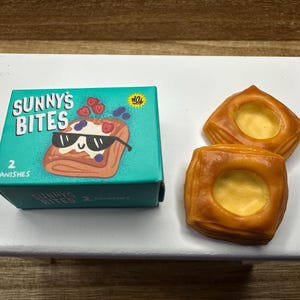 Pode incluir: Uma caixa turquesa de "Sunny's Bites" dinamarqueses com um pastel de desenho animado usando óculos de sol. Dois dinamarqueses castanhos dourados com um recheio cremoso estão ao lado da caixa. A caixa diz "2 Danishes".