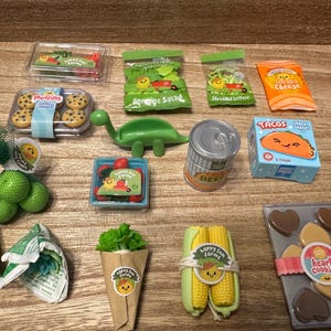 Pode incluir: Uma coleção de alimentos de brinquedo em miniatura, incluindo muffins, alface, queijo, tacos e biscoitos. Os itens estão dispostos em uma superfície de madeira, alguns embalados em plástico transparente ou papel. Há também um brinquedo de dinossauro verde.