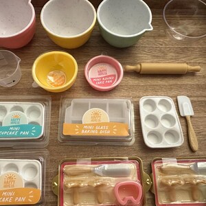 Pode incluir: Uma coleção de mini ferramentas e acessórios de panificação, incluindo tigelas em rosa, amarelo e verde, um rolo, xícaras de medição e formas de cupcake. O conjunto também inclui uma mini assadeira de vidro e uma espátula.