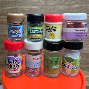 Pode incluir: Uma coleção de pequenos frascos de especiarias com vários rótulos, incluindo "Peppercorn", "Tzatziki" e "Horseradish". Os frascos têm tampas de cores diferentes e contêm ingredientes como cerejas e nonpareils arco-íris.